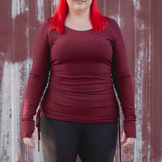 Person wearing a maroon long-sleeve top, black leggings, and brown boots standing in front of a weathered red wall.