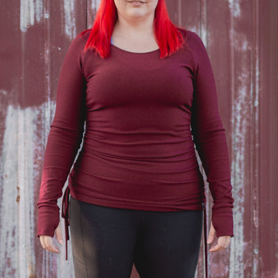 Person wearing a maroon long-sleeve top, black leggings, and brown boots standing in front of a weathered red wall.