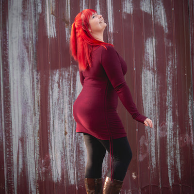 Person with red hair wearing a burgundy dress and brown boots standing against a rustic metal wall.