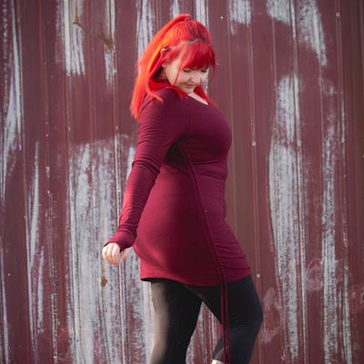 Person with red hair wearing a red dress and brown boots standing in front of a weathered red wall.