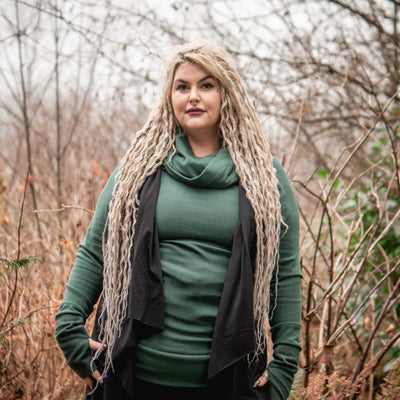 Woman with long blonde hair wearing a green Goddess sweater and black Gemini Vest standing in a forest.